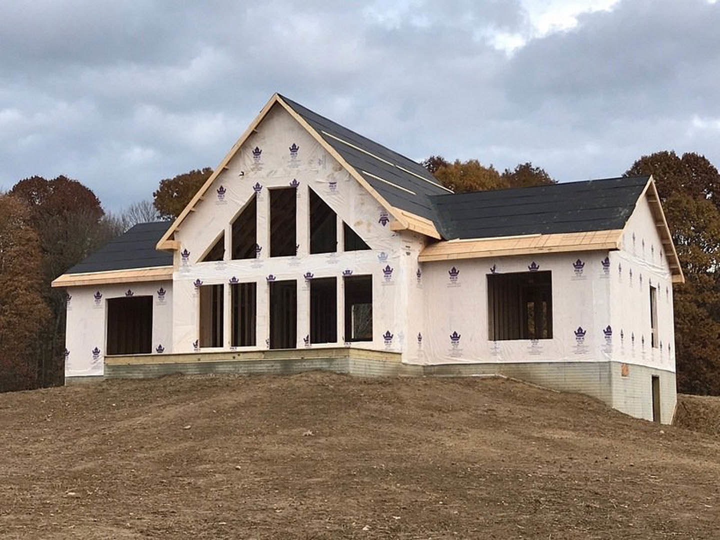 Partially built farmhouse with black roof, white exterior walls, and dirt yard, surrounded by trees under a cloudy sky