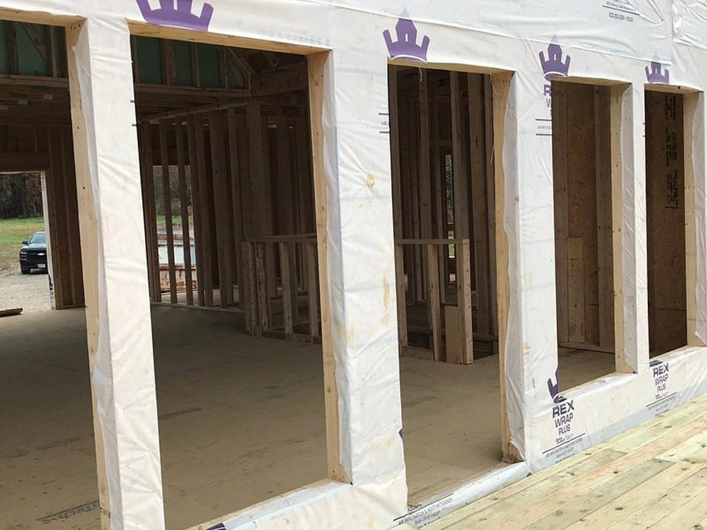 Modern home under construction featuring white exterior walls, large windows, and construction materials scattered on the ground; partial view of a parked car in the foreground.