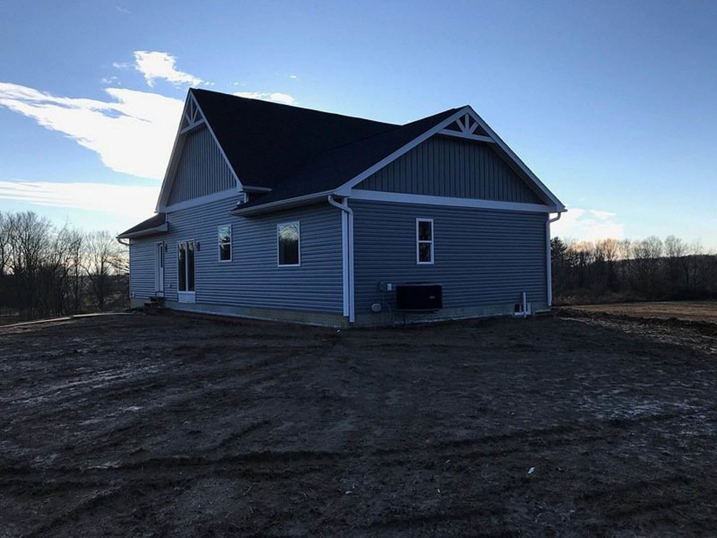 Partially built house with black roof and light siding, surrounded by dirt field with tire tracks, distant trees under cloudy sky