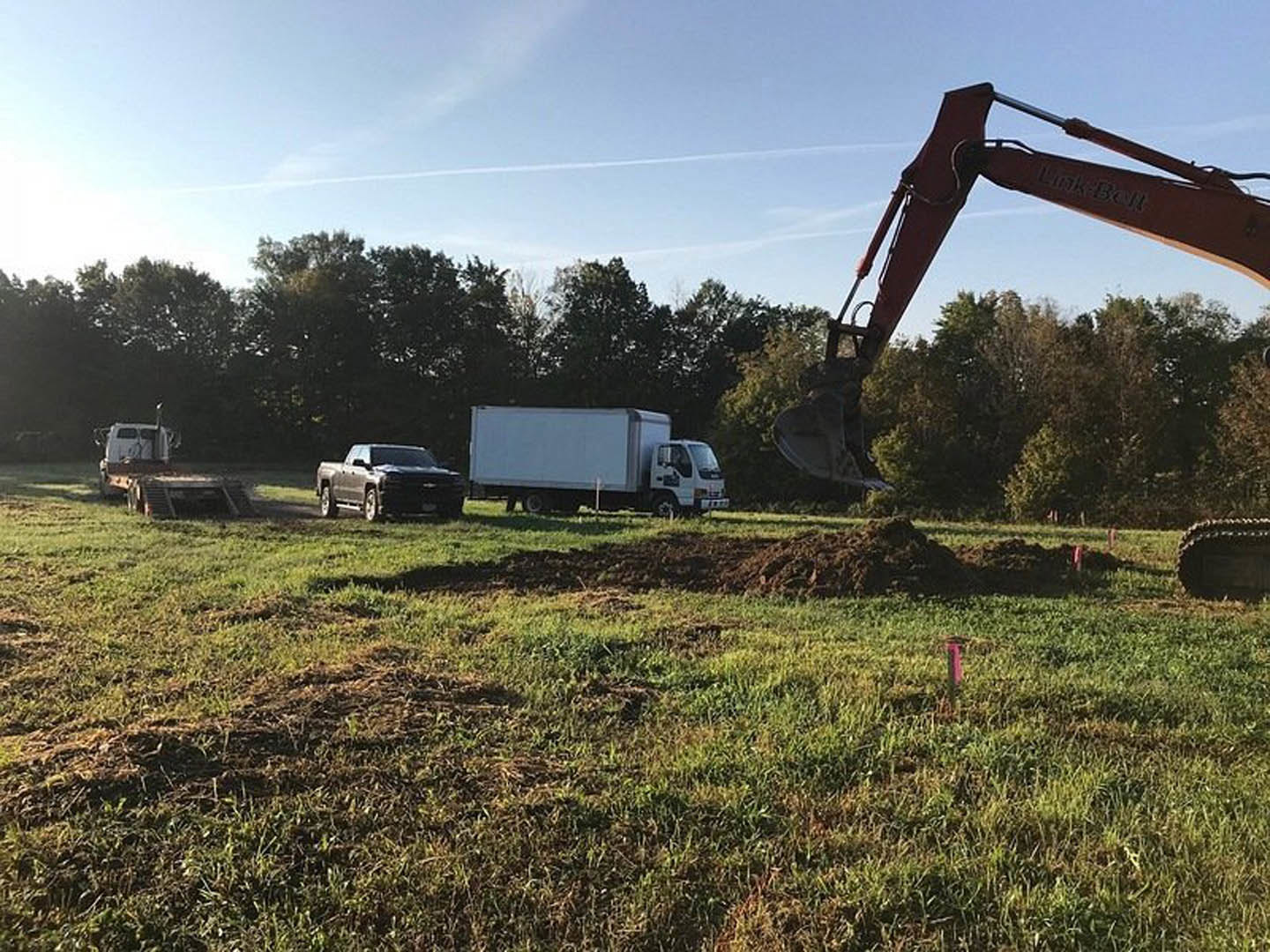 Red excavator digging foundation hole on grassy construction site, white truck parked nearby, dirt piles and scattered vehicles visible under open sky.