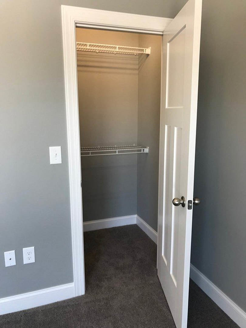 Open closet door revealing white metal shelf with black square pattern, white wall outlet, light switch, grey carpeted floor, and light grey walls.