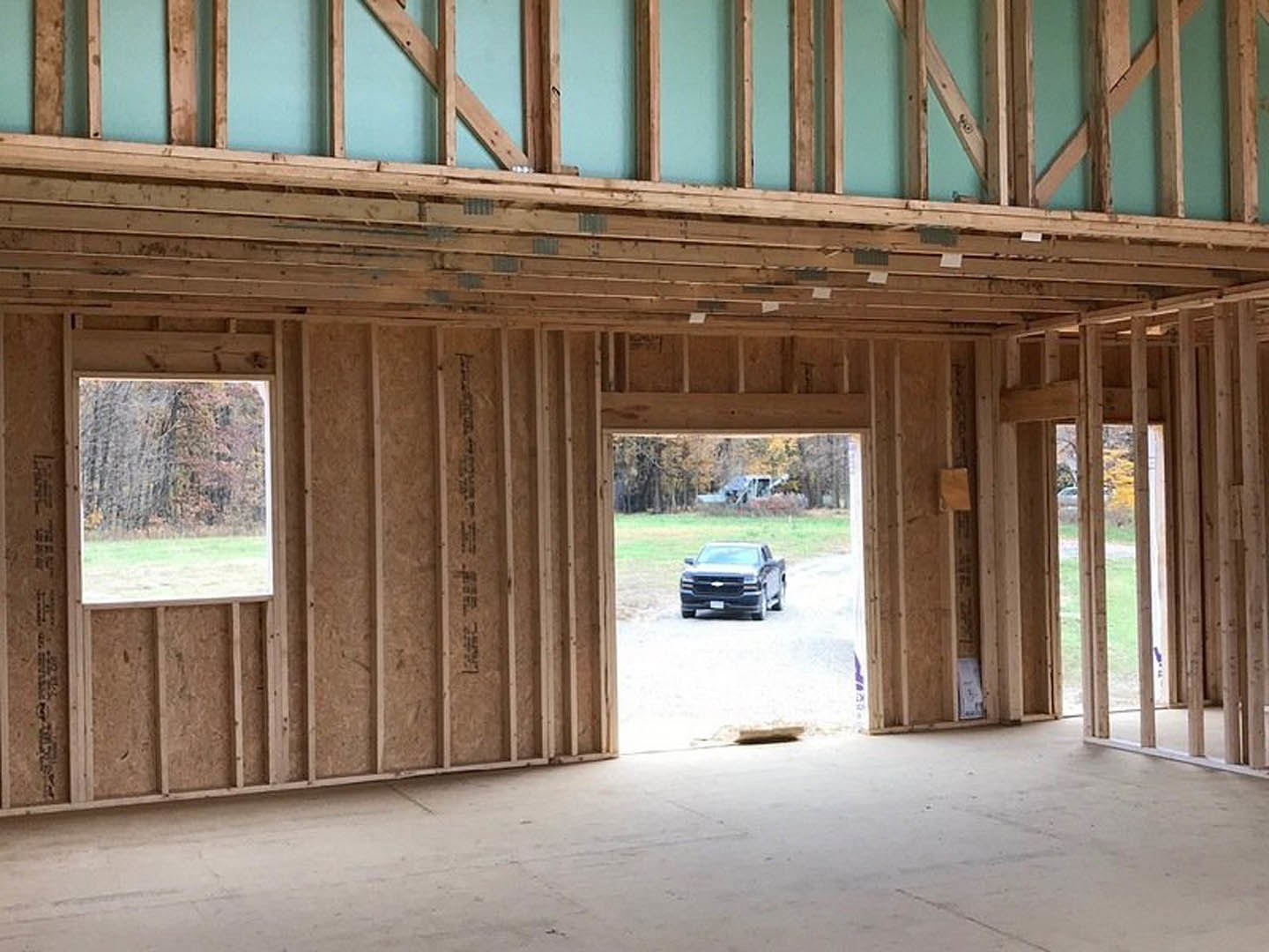 Open-concept interior with exposed wood beams, unfinished concrete floor, large window overlooking trees, and a black truck parked on a dirt road outside.