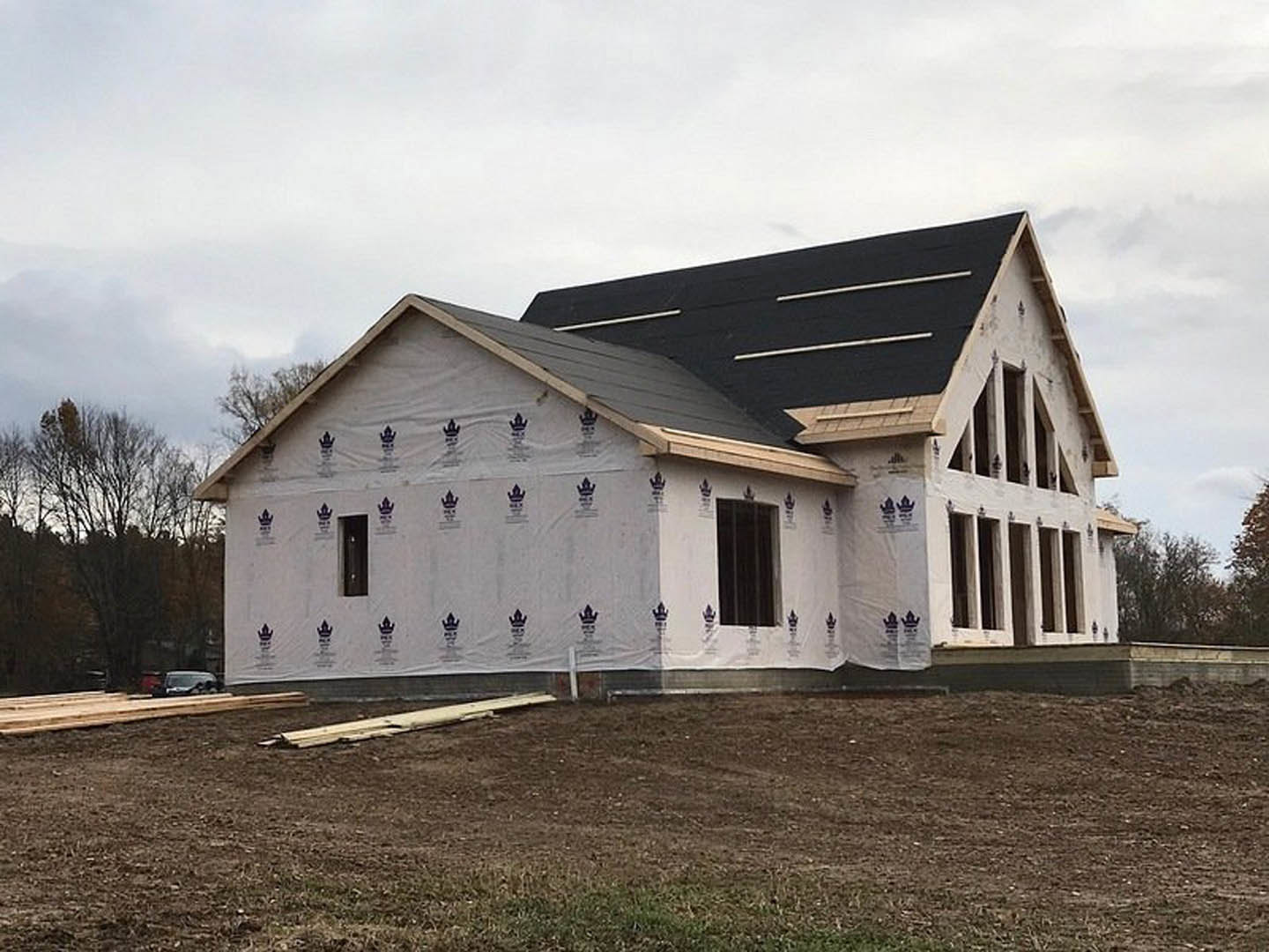 Two-story house under construction with black roof, white siding covered in protective plastic, large windows, dirt yard, white fence, leafless tree, and cloudy sky