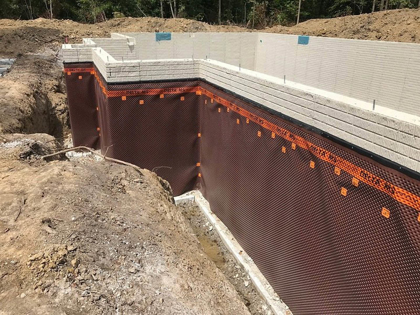 Concrete house foundation with orange mesh reinforcement, surrounded by dirt and construction materials, tree visible in background