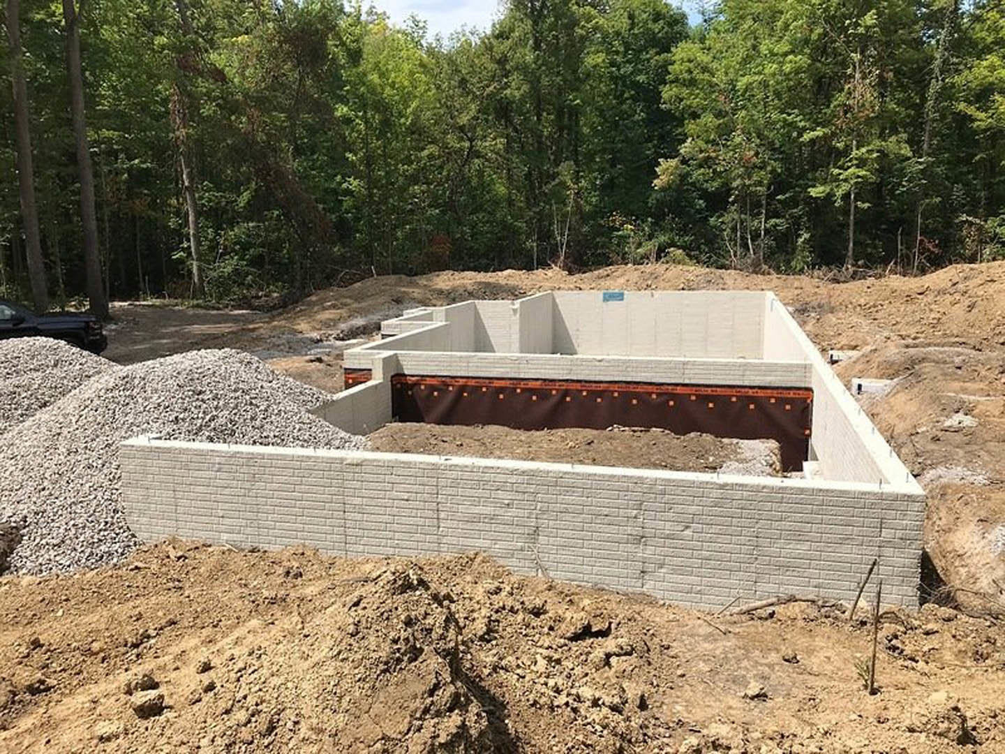 Dirt-covered building site bordered by trees, house foundation and brick wall visible, pile of gravel near construction area