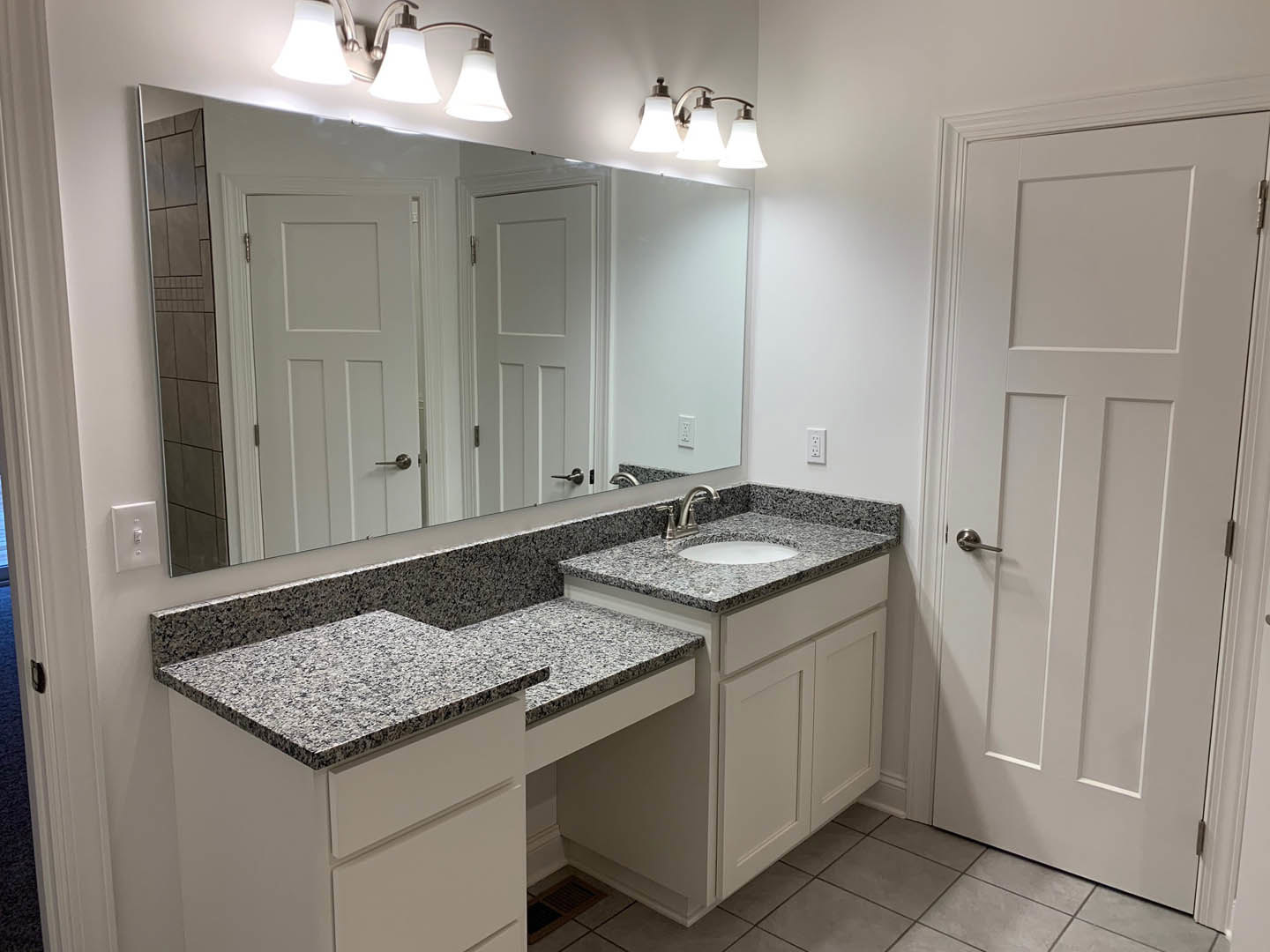 Bathroom with double vanity featuring white sinks, marble countertop, large framed mirror, chrome faucets, white cabinetry, silver hardware, and white door with silver handle.