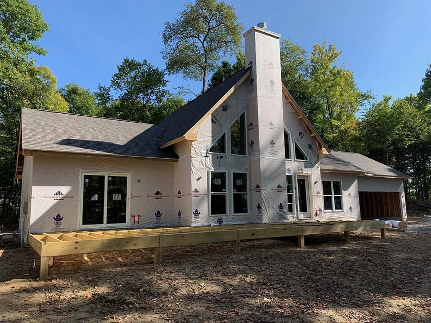 Framed house under construction with wooden platform, white sheeting, chimney, and surrounding trees in background