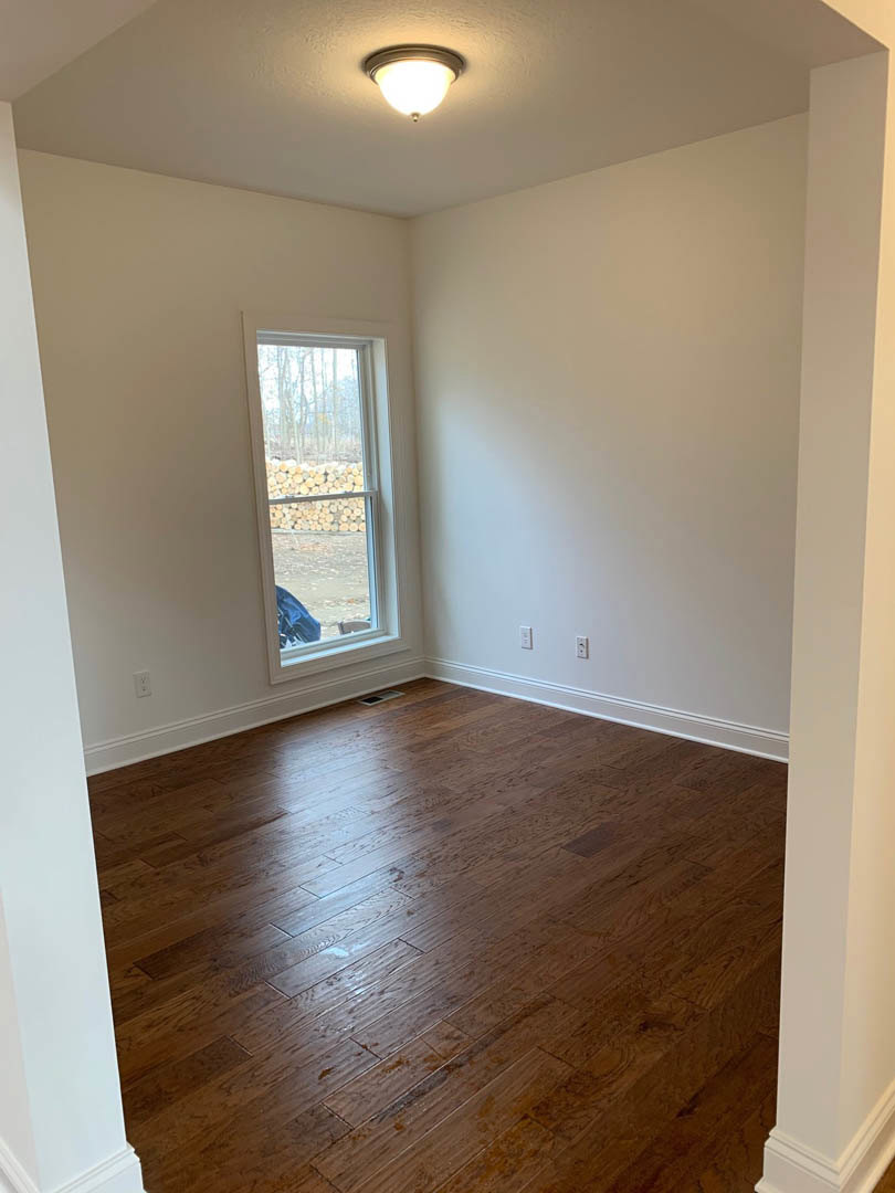 Hardwood floor with natural wood grain, white plaster walls, large window framed in white, ceiling-mounted light fixture, stack of firewood near the window, room corner visible