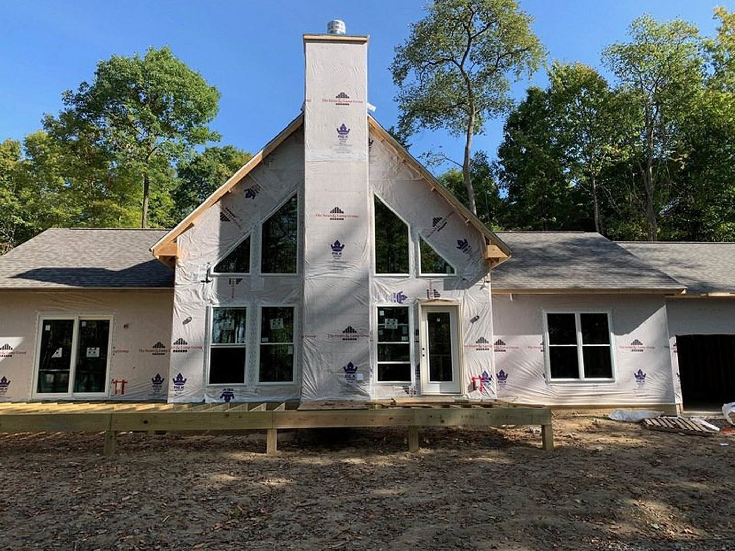 Framed house under construction with plastic sheeting, exposed wooden deck, windows with signage, surrounded by trees