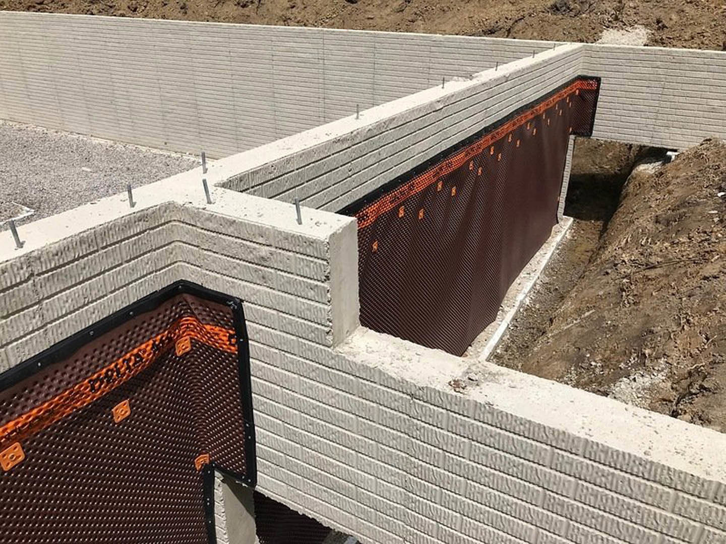 Brick exterior wall under construction with black and orange mesh mat covering, adjacent to a ground trench