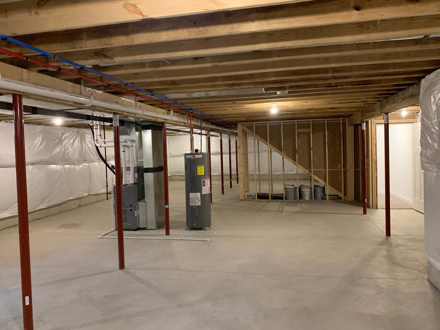 Basement under construction featuring exposed wood ceiling beams, visible blue electrical wires, metal support pole, concrete floor, staircase, and a metal junction box.