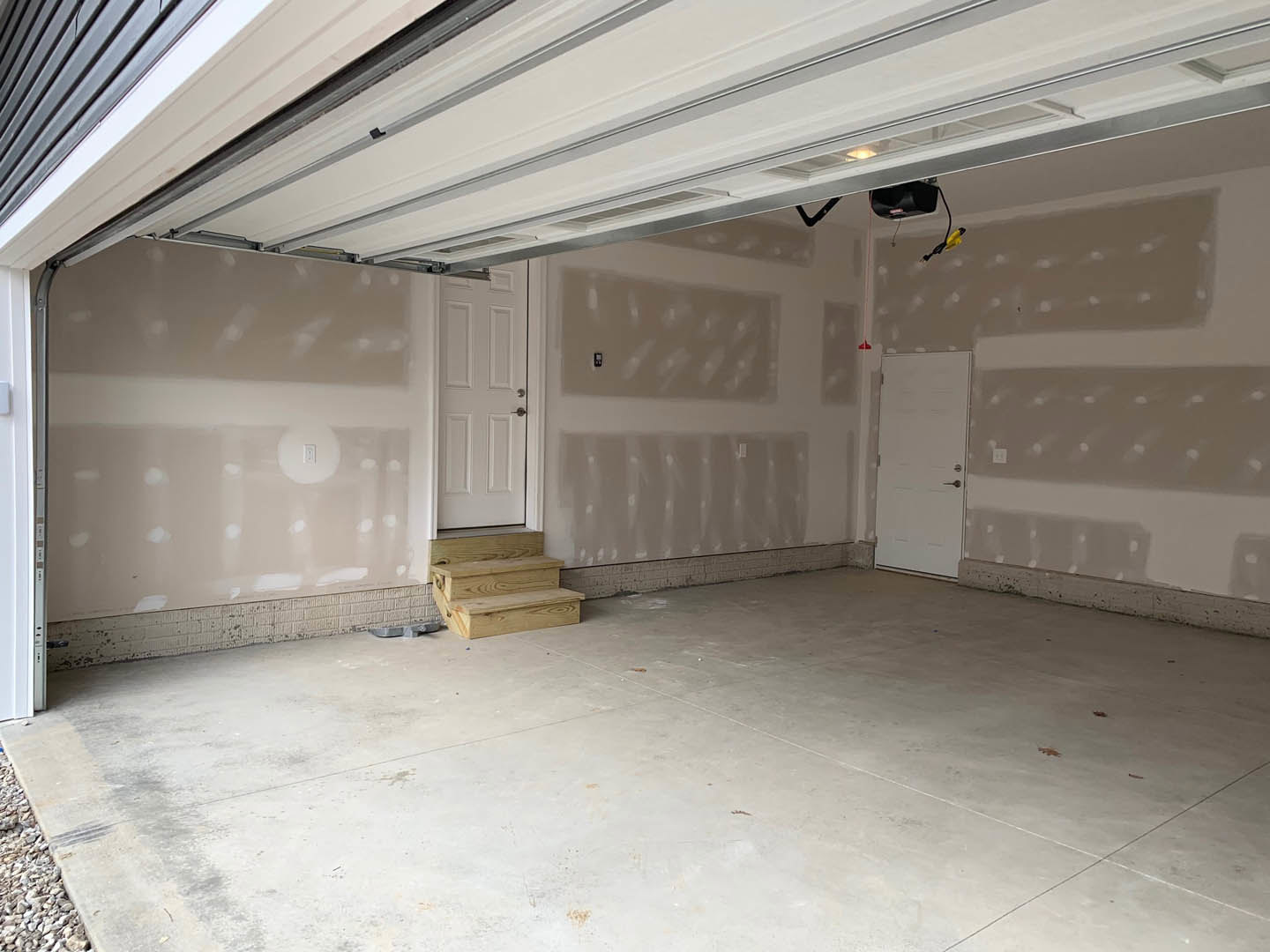 Garage interior with two white paneled doors, concrete floor, wooden steps leading to a raised entry, plaster walls, and exposed ceiling insulation.