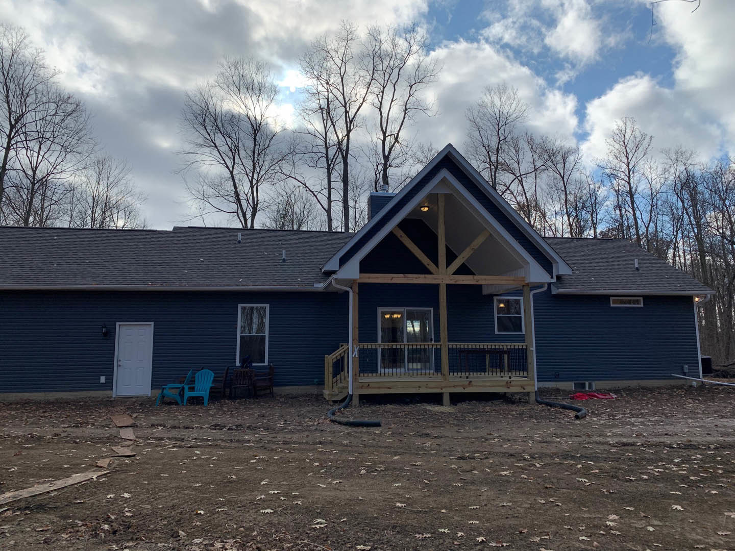 Two-story home with light siding, covered front porch, wooden deck with railing, white door with black shutters, blue plastic chairs beside blue wall, white-framed window