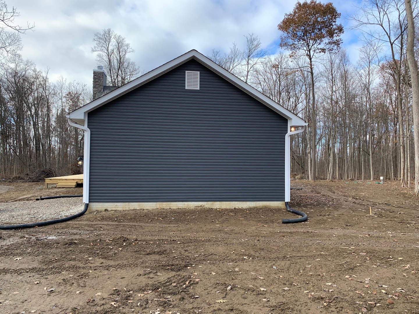 White vent mounted on exterior wall beside brick chimney, hose lying on dirt lot, unfinished wooden stairs leading to entry, metal shed in background, scattered trees under cloudy
