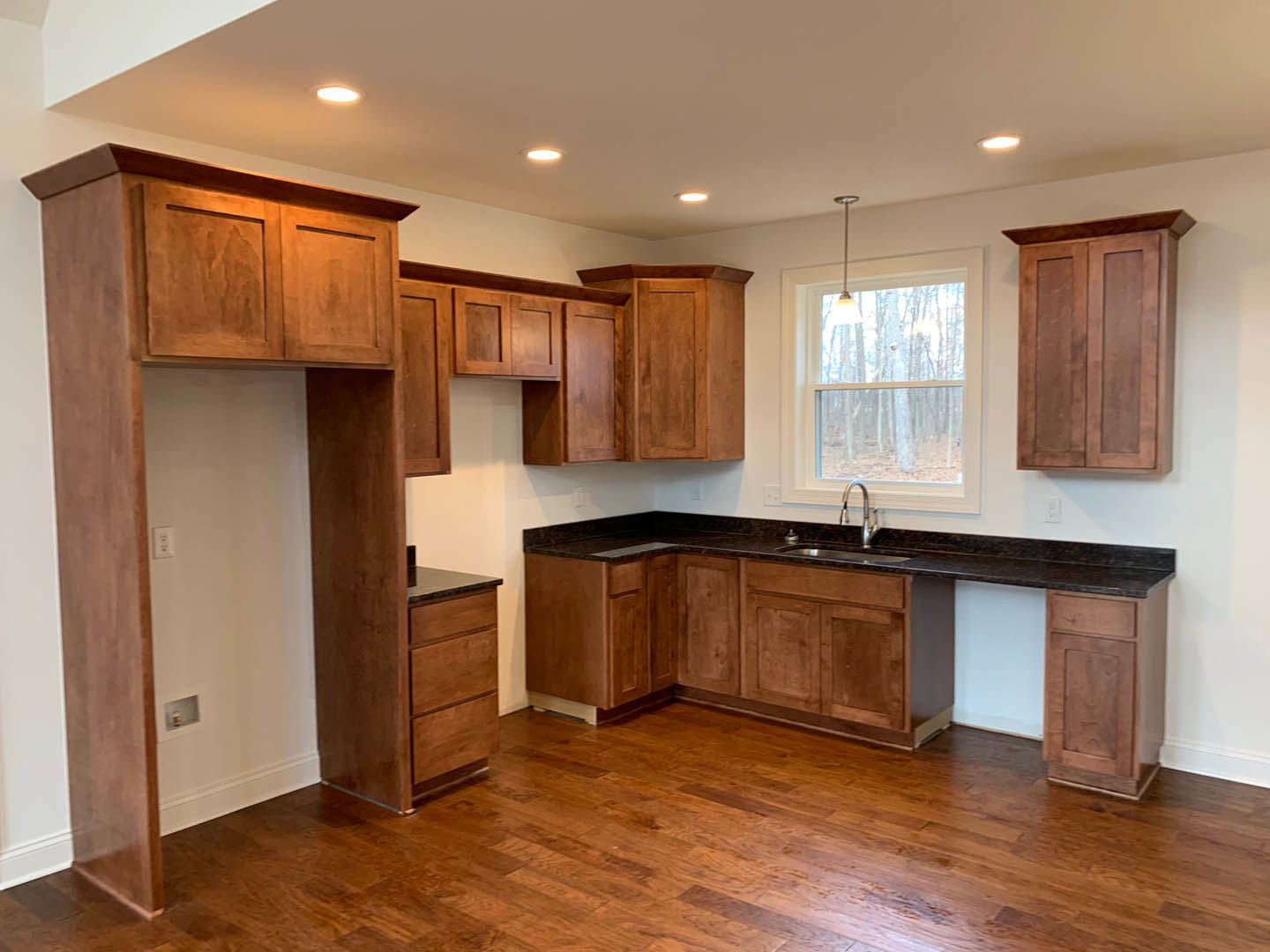 Kitchen with natural wood cabinets, wide window overlooking forest, stainless steel sink, wood flooring, stone countertop, and modern light fixture.