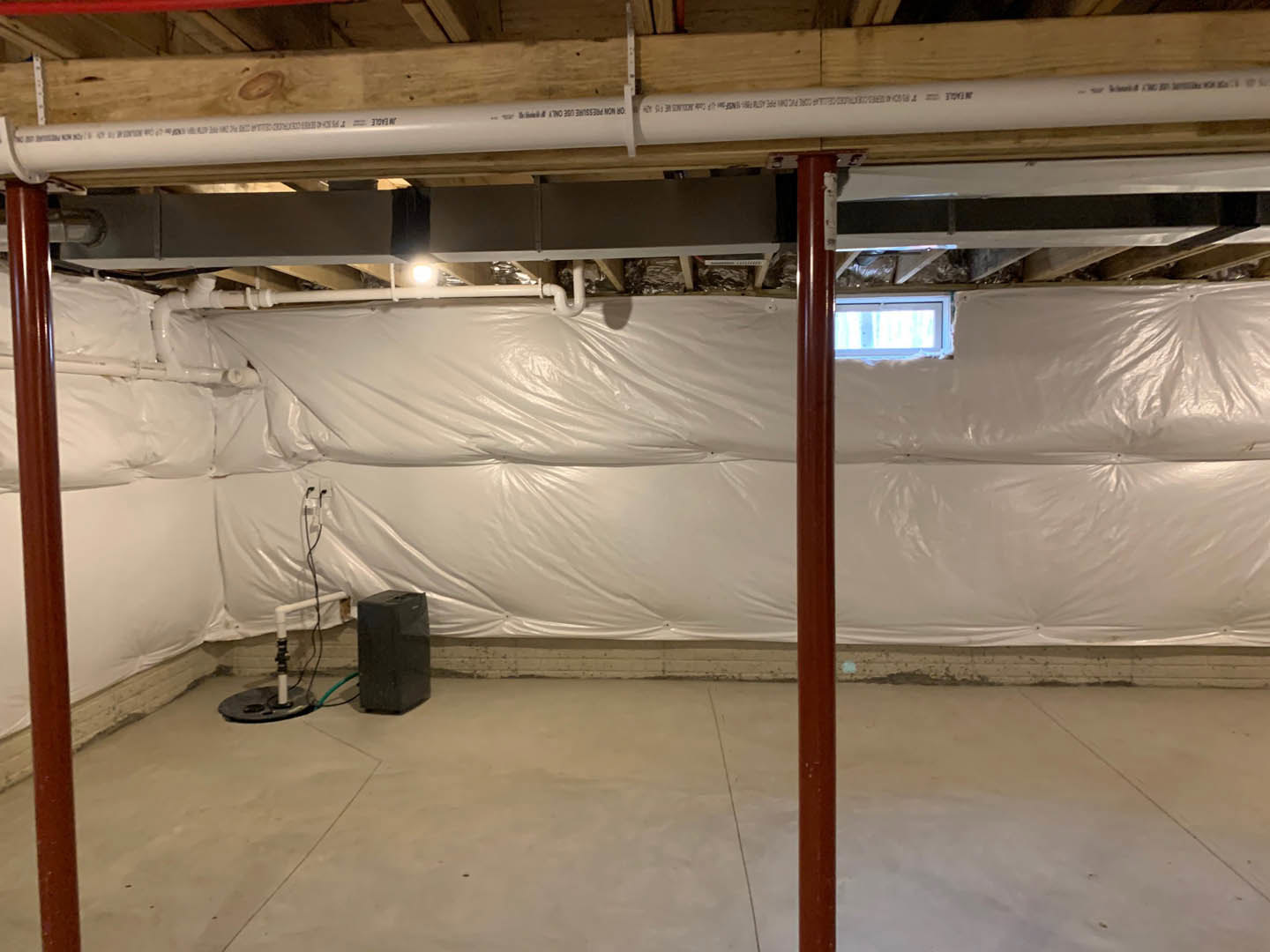 Basement with plastic sheeting covering walls and ceiling, exposed beams, concrete floor, white pipe with black text, hose attachment, blurry window in background.