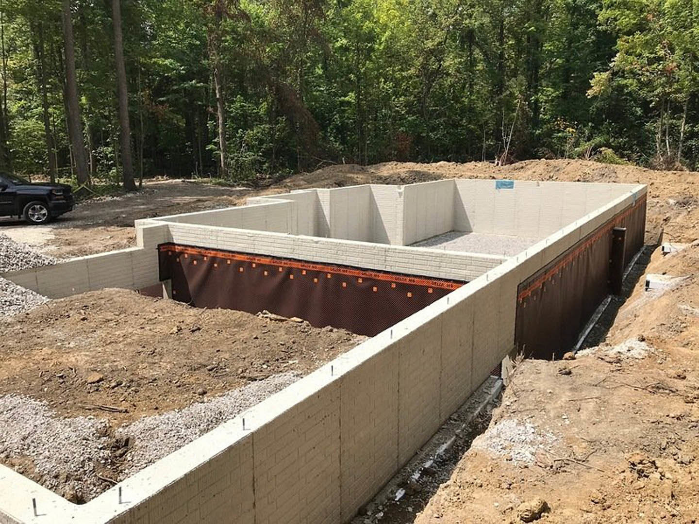 Concrete foundation under construction surrounded by tall trees, red and white formwork panels, black pickup truck parked nearby, dirt path leading through wooded area