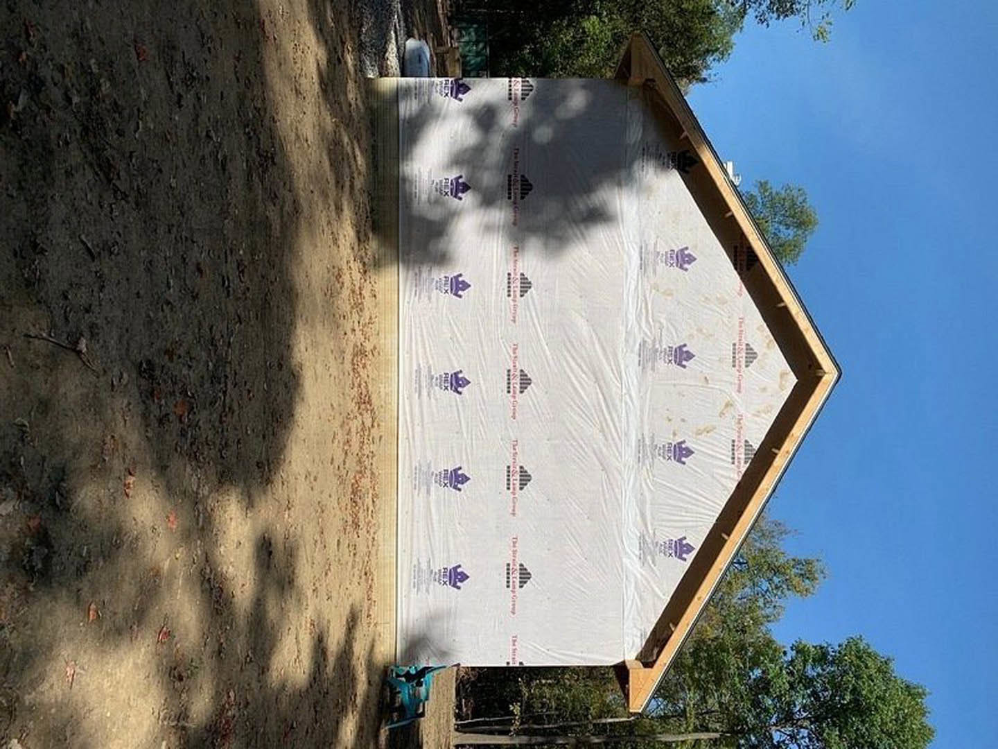 Partially built house with white sheet covering exterior, triangular roof, tree shadows on ground, green trees in background, blue and black construction materials visible