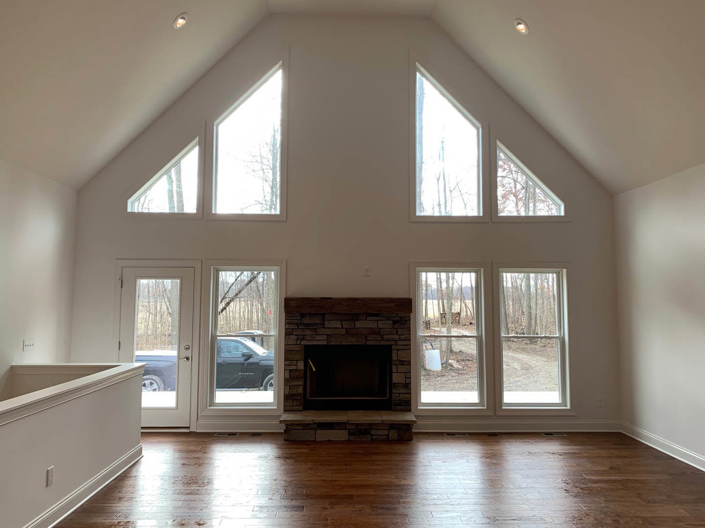 Living room with wood-framed fireplace, large windows, hardwood flooring, and white walls; man standing near fireplace, door with doorknob visible, car parked outside.