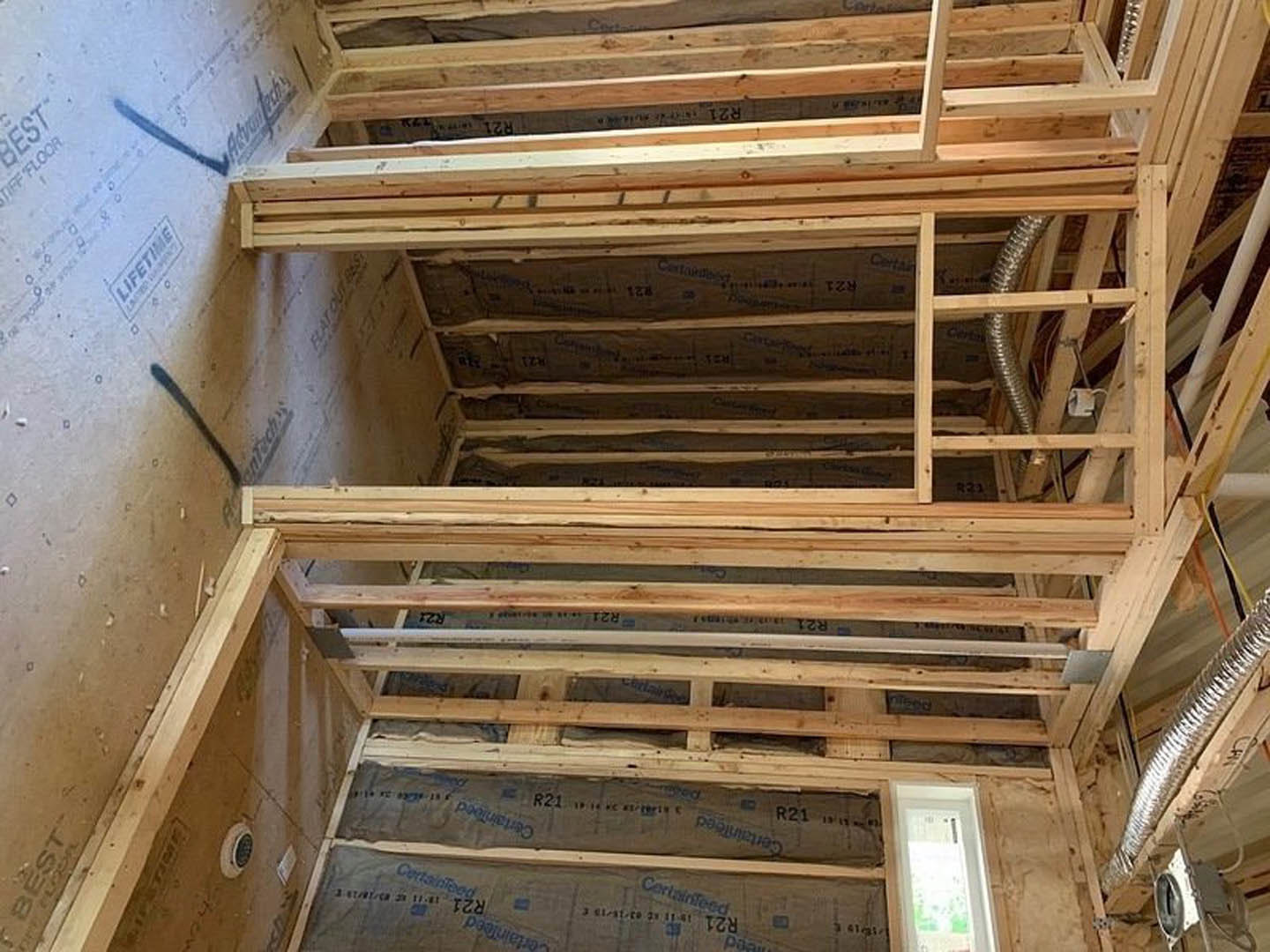 Room under construction with exposed wood framing, white-framed window, metal ductwork, and numbered wooden electrical box.