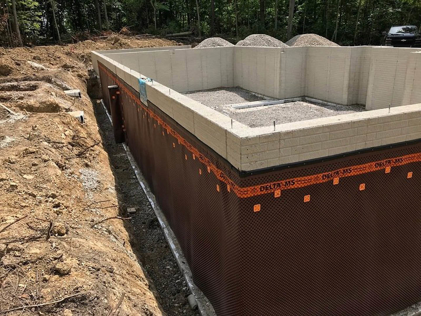Concrete foundation slab surrounded by gravel and dirt, bordered by a metal fence, with trees and plants visible in the background.