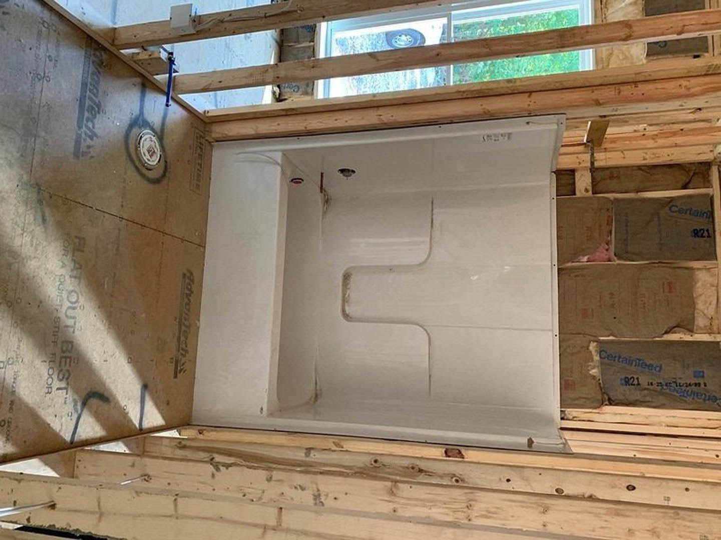 White bathtub with exposed wooden framing, cardboard box with blue writing on unfinished floor, visible floor drain, surrounding walls with construction materials and insulation.