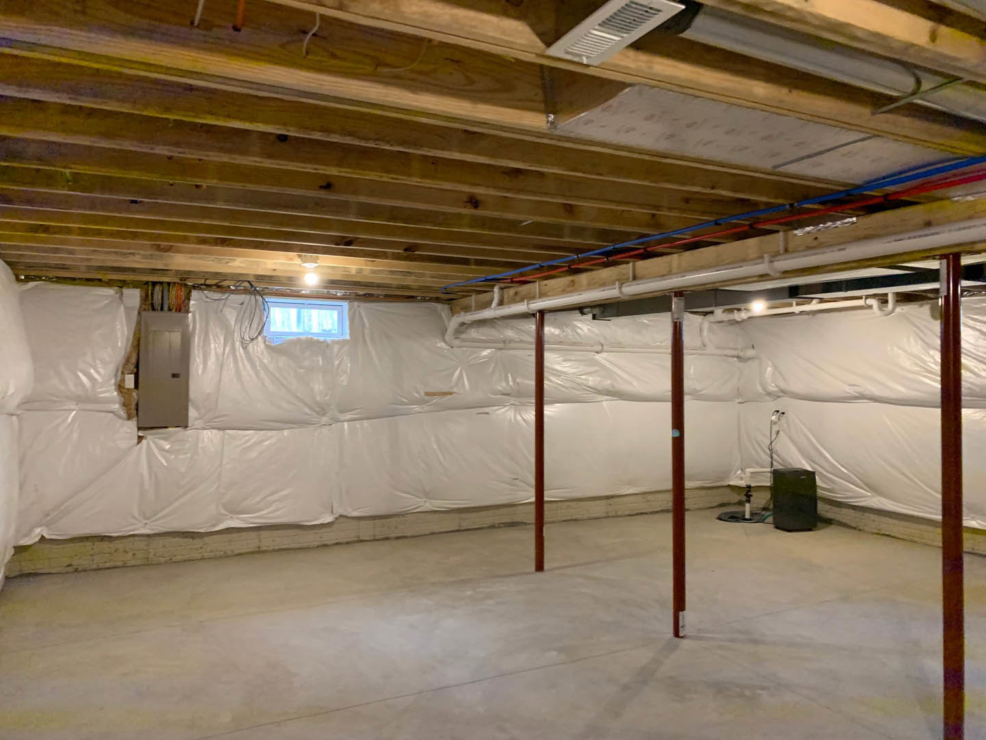 Basement room with concrete floor, exposed ceiling beams, and white plastic coverings draped along walls and insulation; white box with black square sits near vent, plastic bags