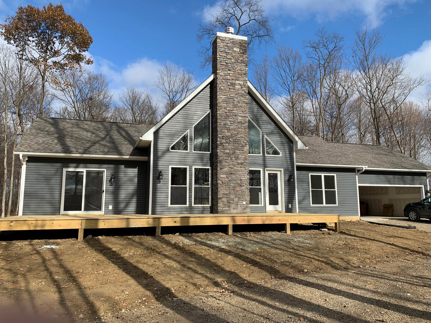 Two-story cottage-style home with brick chimney, white siding, large windows, and a covered porch with wooden decking, surrounded by dirt landscaping and mature trees.
