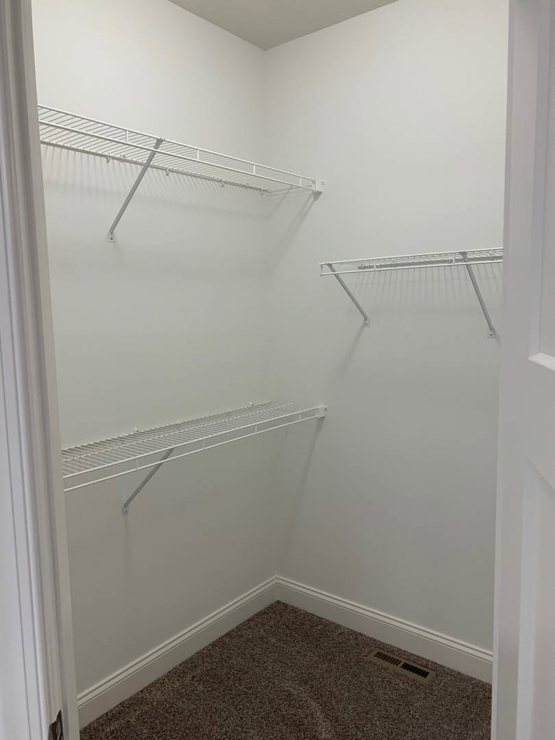 White closet interior with multiple metal shelves and hanging bars, carpeted floor, wall vent, and smooth white plaster walls.