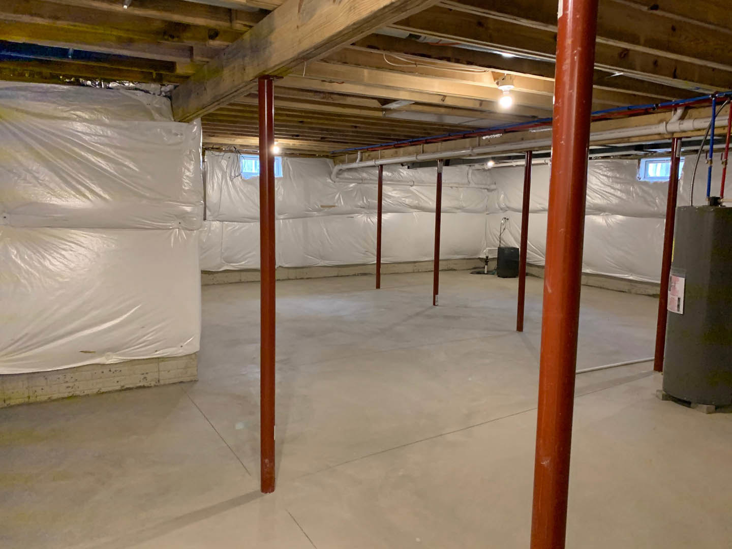 Basement with exposed wooden ceiling beams, white plastic sheeting covering walls and surfaces, red steel support pole wrapped in white, window showing blue sky, scattered white