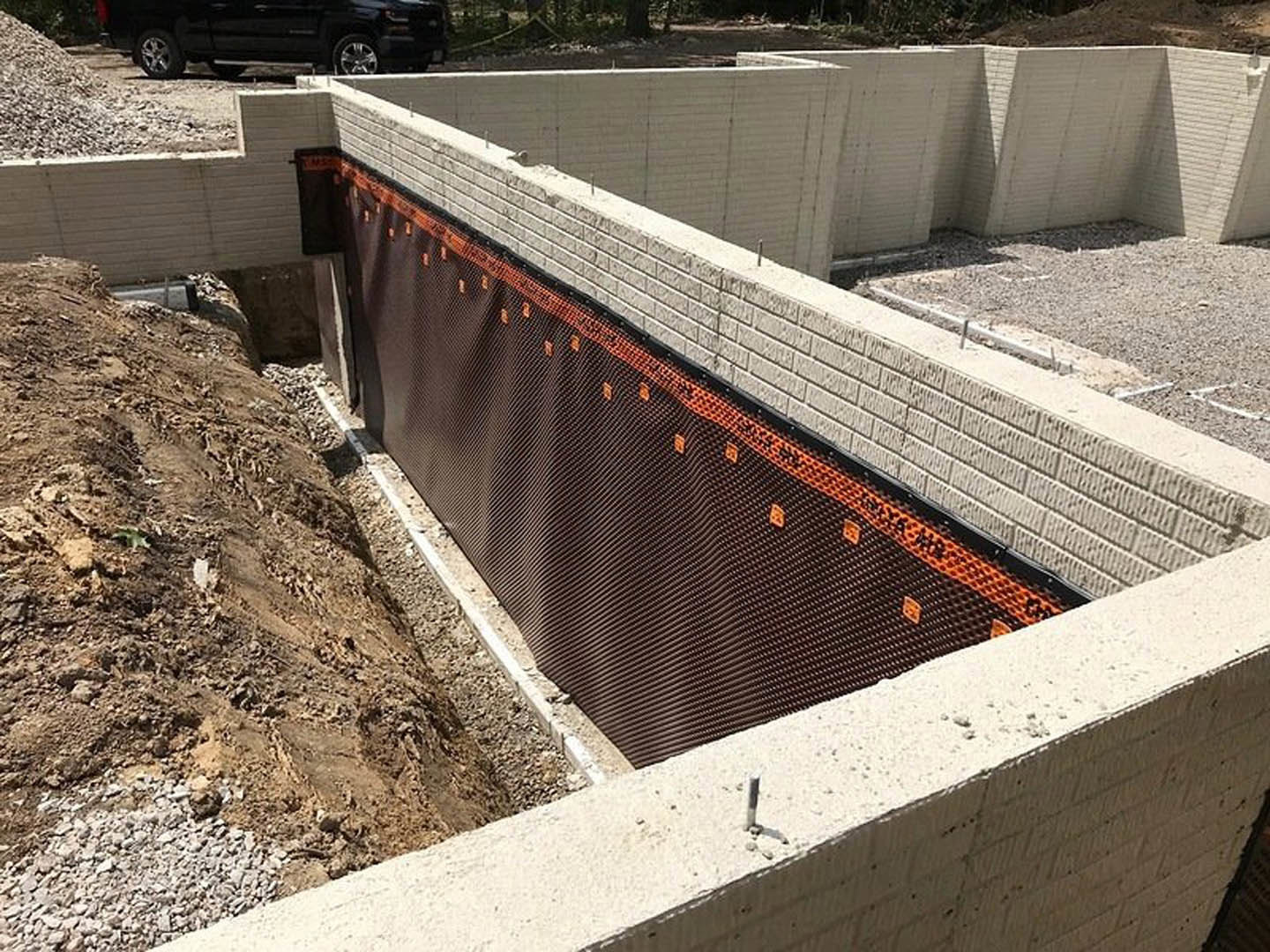 Concrete foundation wall with black waterproofing membrane and orange mesh, bordered by dirt and gravel, construction materials visible nearby.