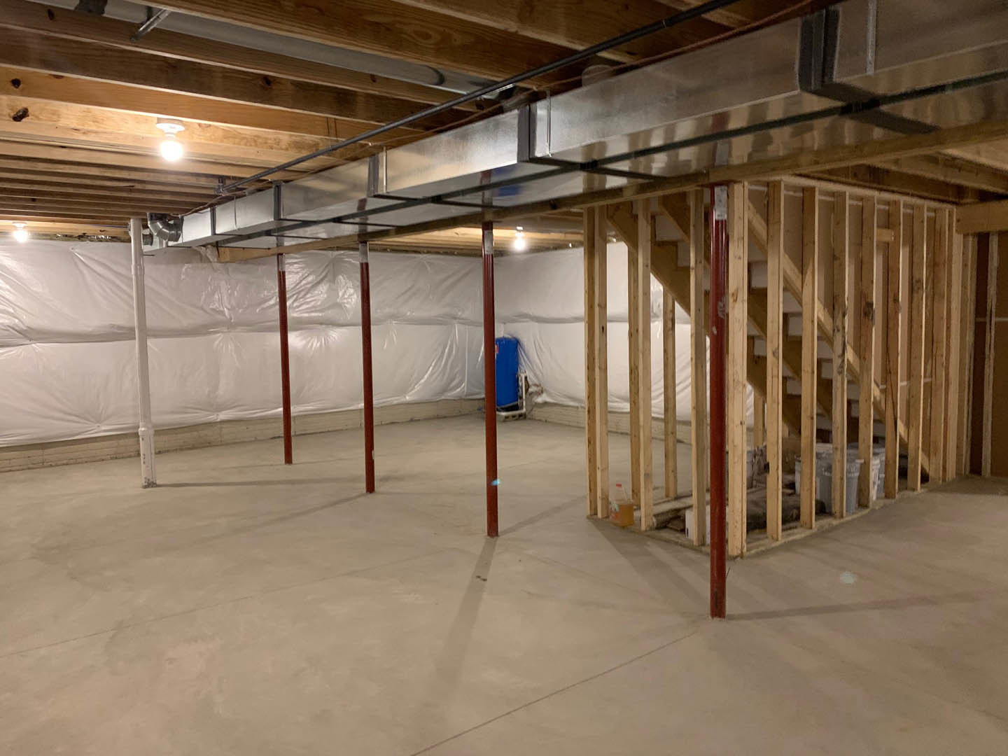 Basement with exposed wood beams and metal pipes on ceiling, concrete floor with red support pole, blue rectangular object with brown frame, white plastic wall covering, small