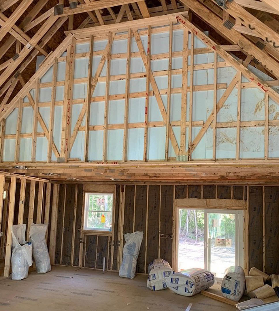 Unfinished room with exposed wooden framing, ceiling beams, and insulation; large windows and glass door provide natural light, construction materials visible on floor.