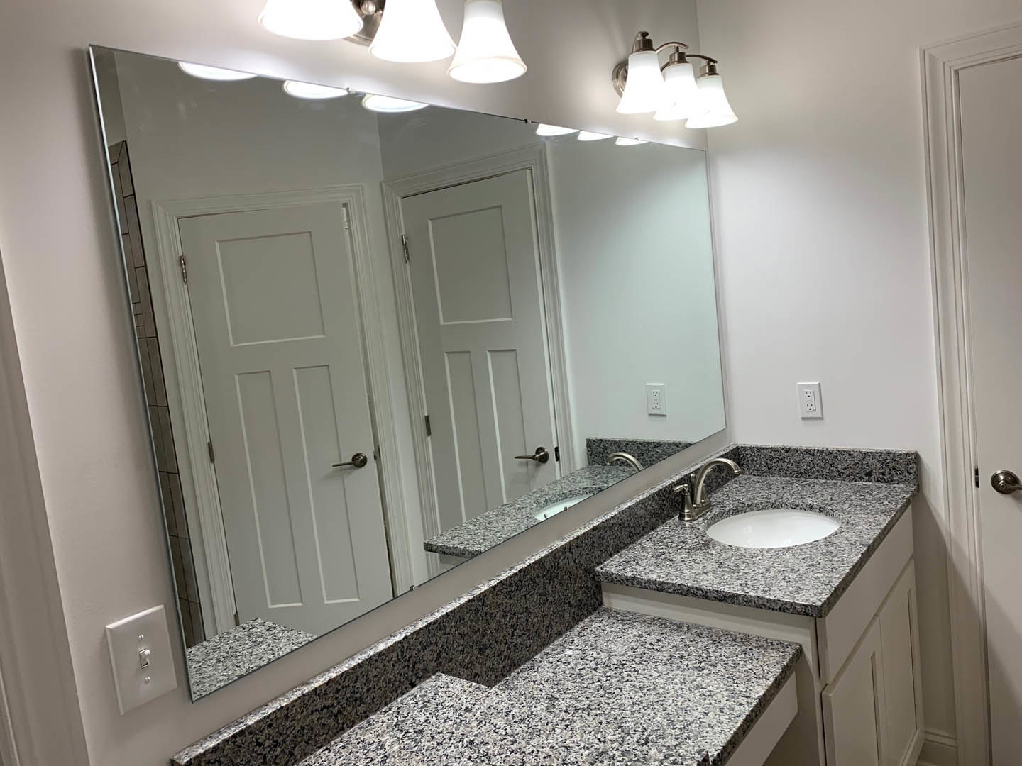 Granite countertop with undermount sink and chrome faucet, large framed mirror above, three-light fixture, white plate on counter, tiled walls