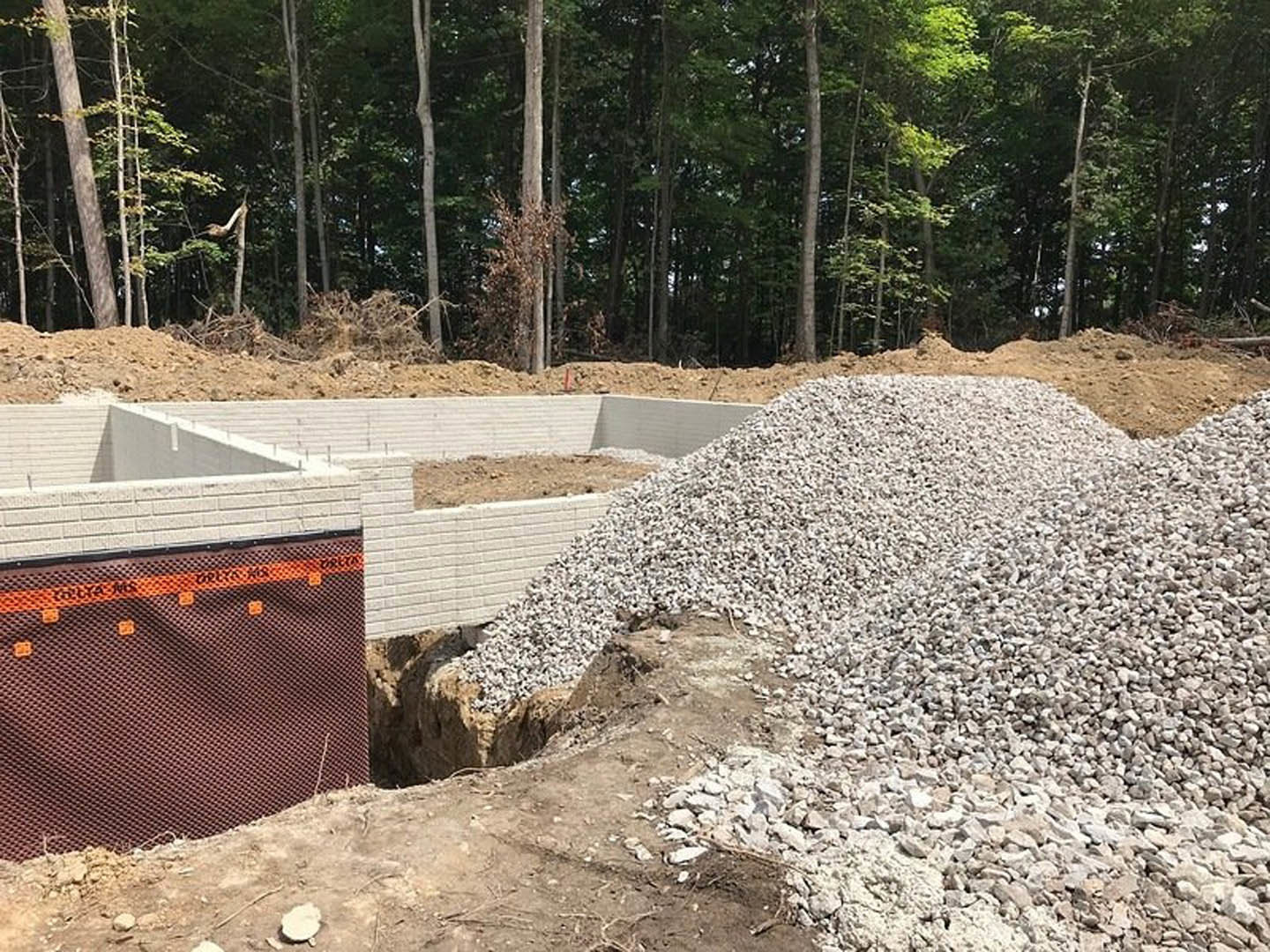 Gravel pile beside poured concrete foundation, brown and orange construction barrier in foreground, forested area with tall trees in background, scattered rocks and dirt on ground