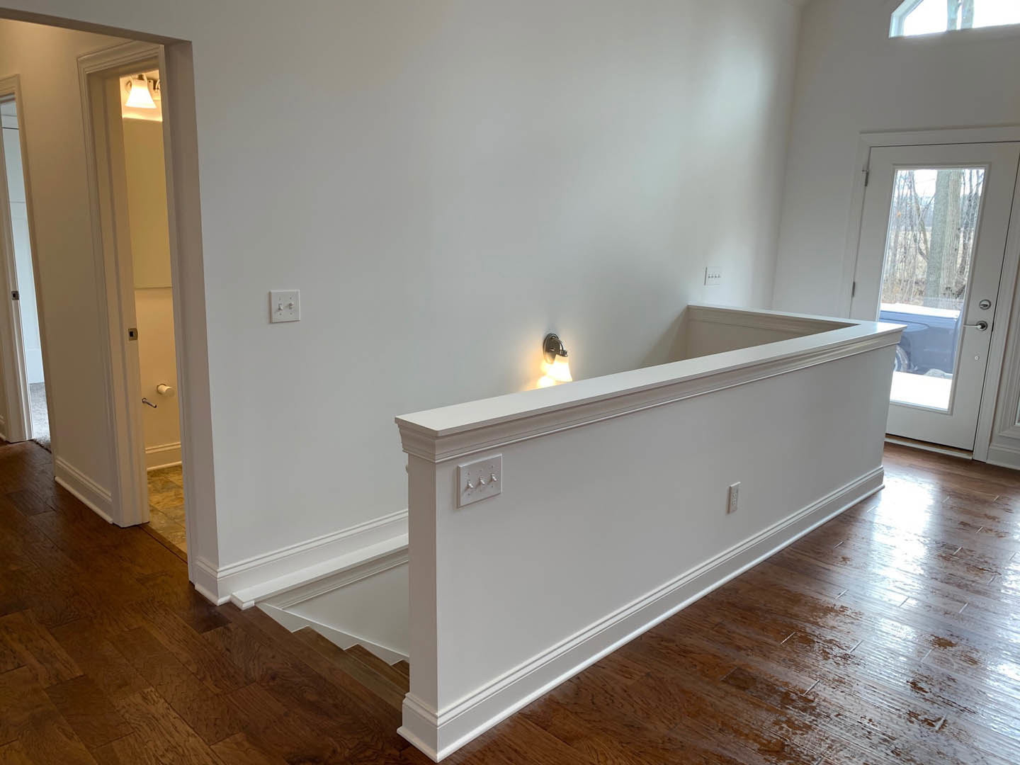 White staircase with wood treads beside a white plaster wall, illuminated by wall-mounted light; light switch and white door with glass window visible, window shows tree outside.