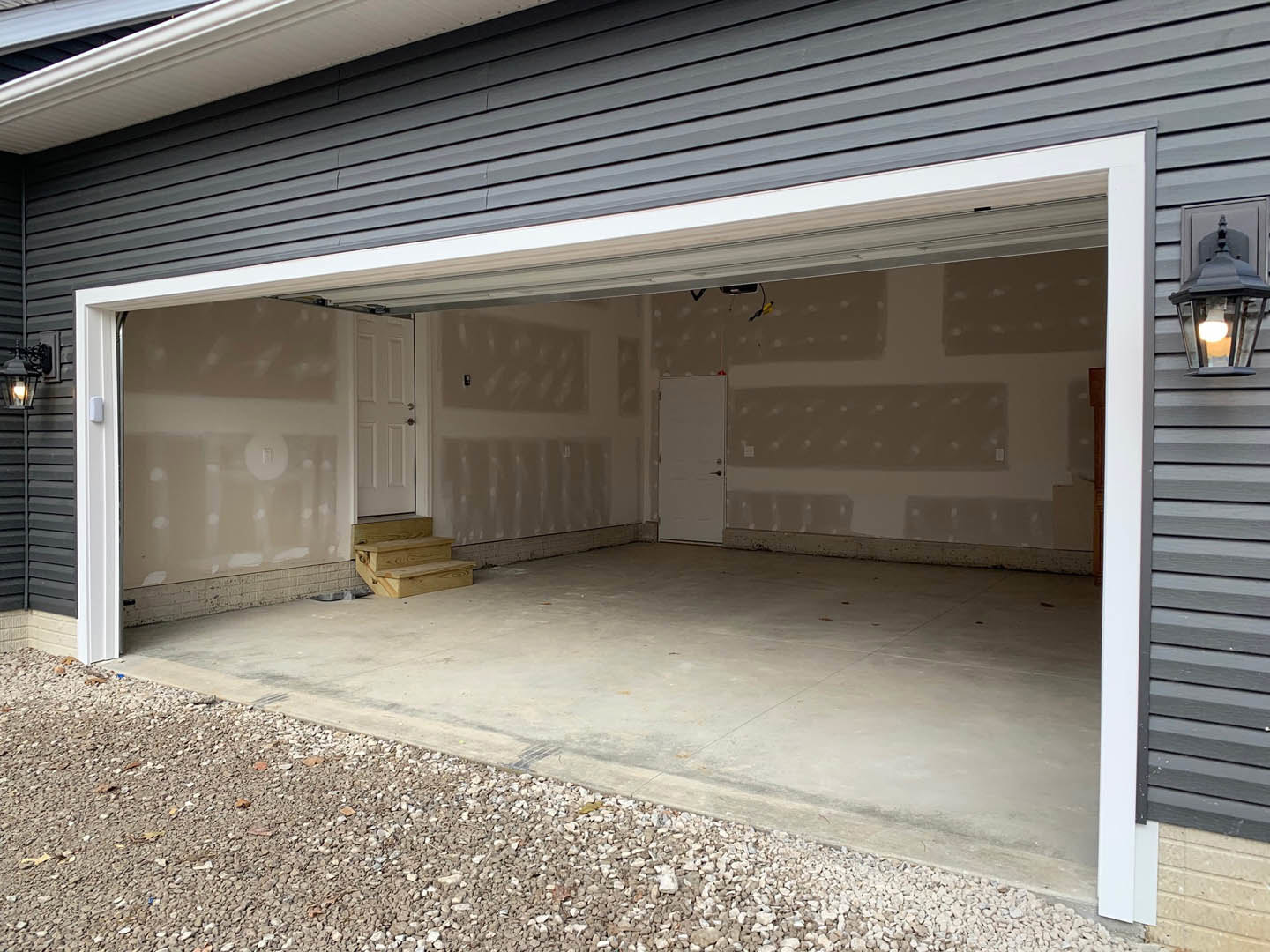 Garage with paneled white door, concrete floor, gravel border, and black handle; adjacent white door and wooden staircase visible inside.