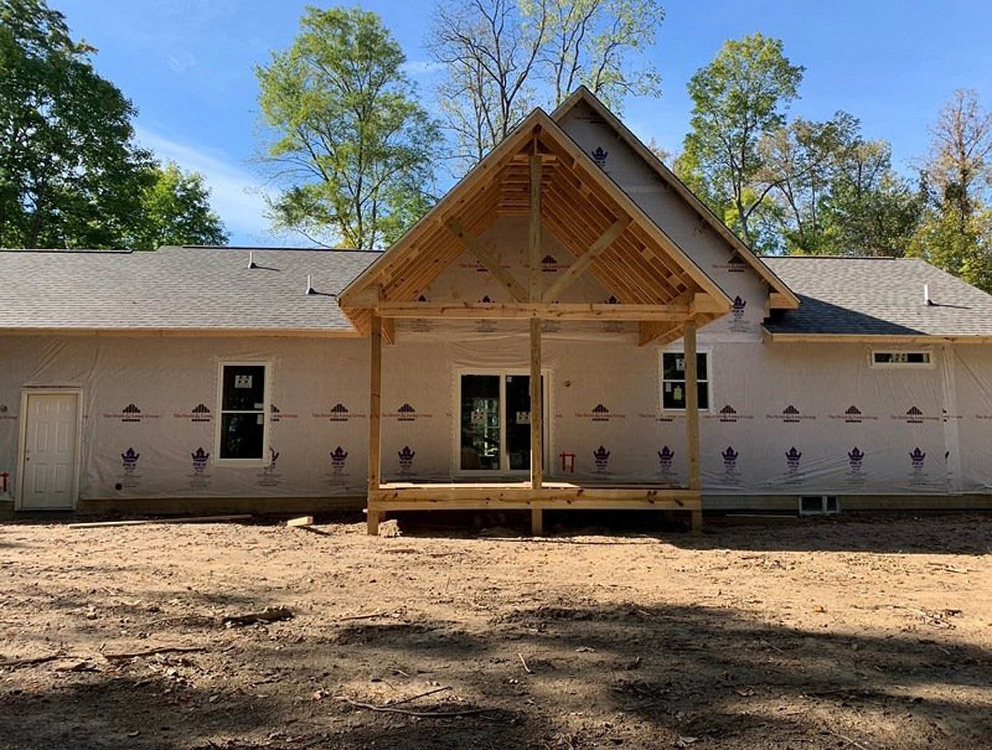 Wood-framed roof atop partially built house, white door with silver doorknob, white-framed window, dirt ground scattered with sticks and branches