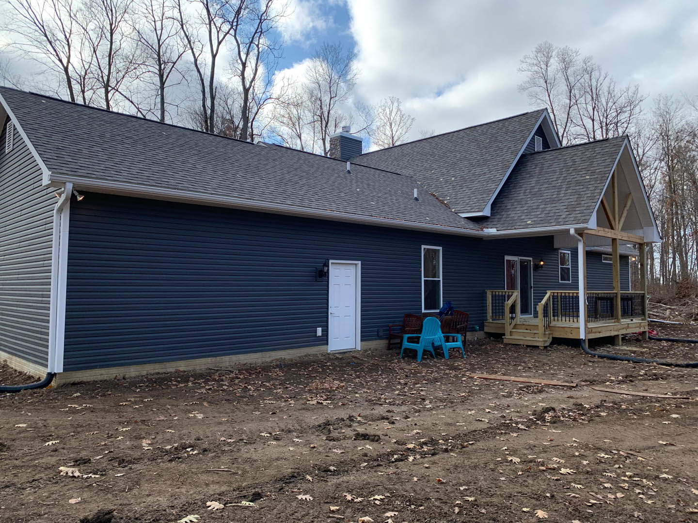 Blue-sided house with white-framed window, white door, covered porch, and attached deck; dirt yard features blue plastic chairs; cloudy sky and trees in background
