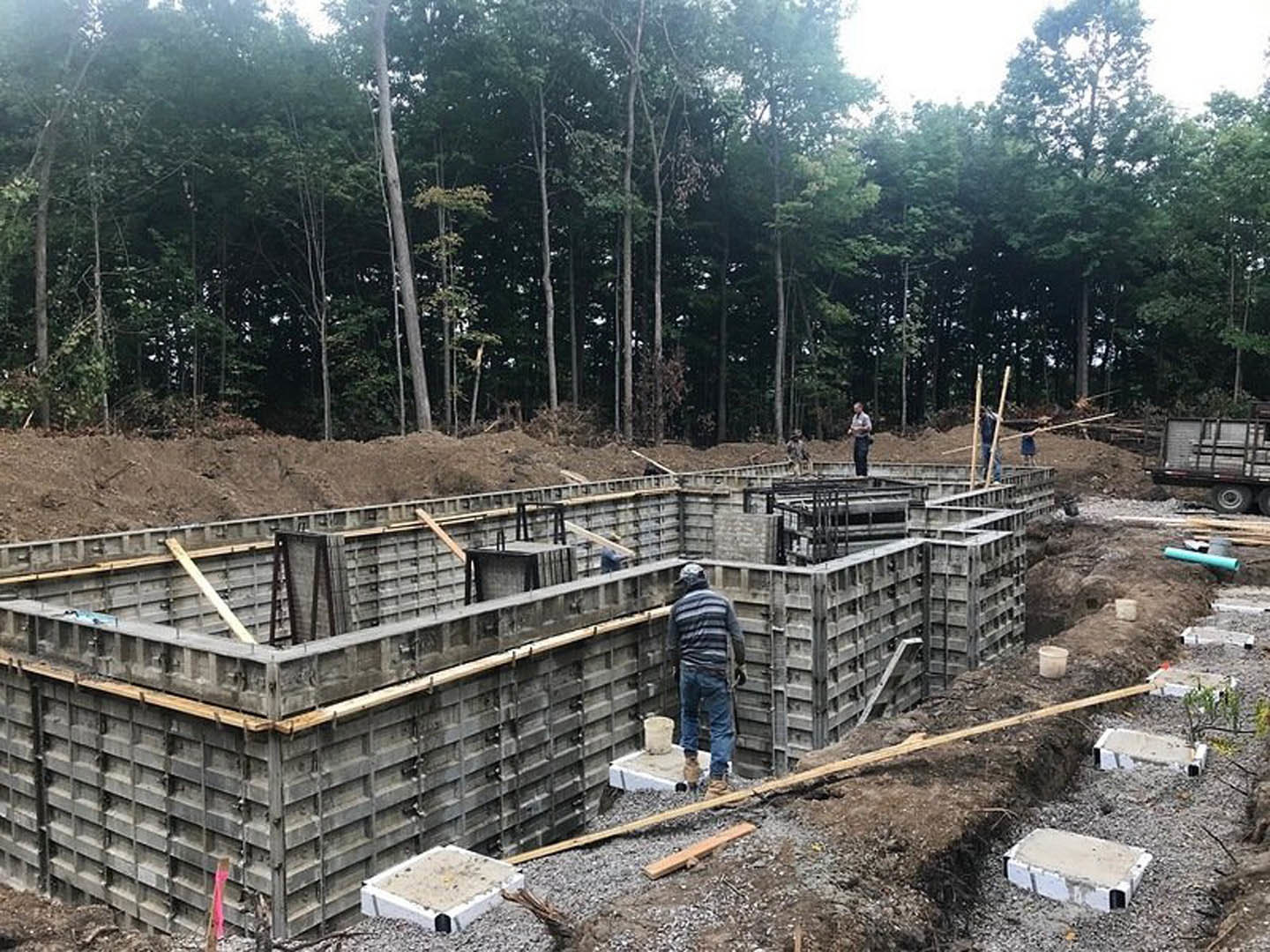 Concrete foundation surrounded by gravel, several workers in casual clothing, metal-framed truck parked nearby, forested area with tall trees in background, white rectangular