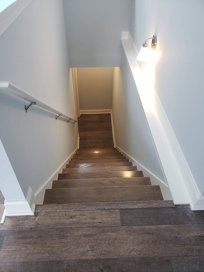 Wood staircase with metal handrail, warm wall sconce lighting, white plaster walls, and natural wood flooring