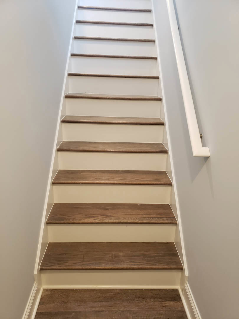 Brown wood staircase with white handrail mounted on adjacent wall, close-up view of wood grain and smooth painted surfaces.