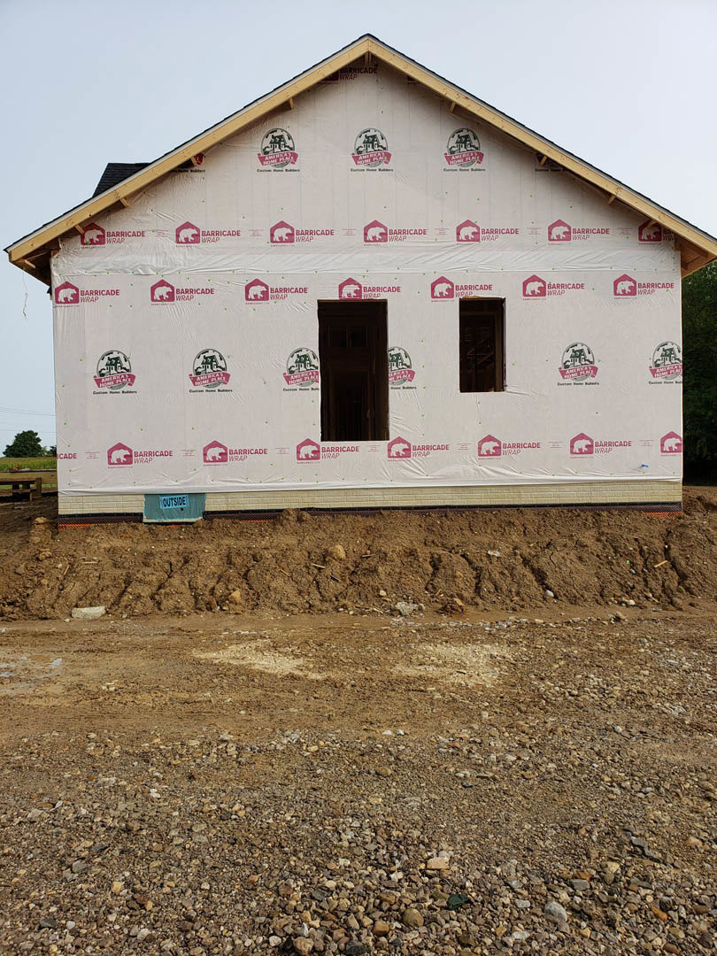 Partially built house with exposed framing, white protective sheets covering exterior walls, dirt mound and rocks in foreground, dark doorway with construction sign, temporary