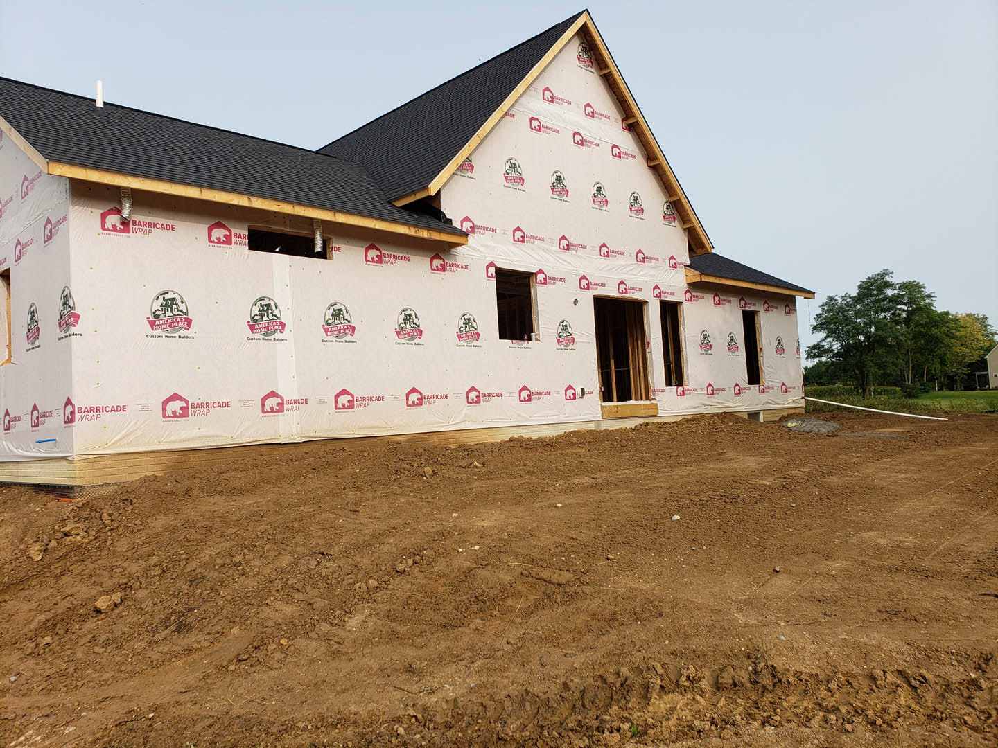 Wood-framed house under construction with black roof, surrounded by dirt and mature trees, white sign visible in foreground