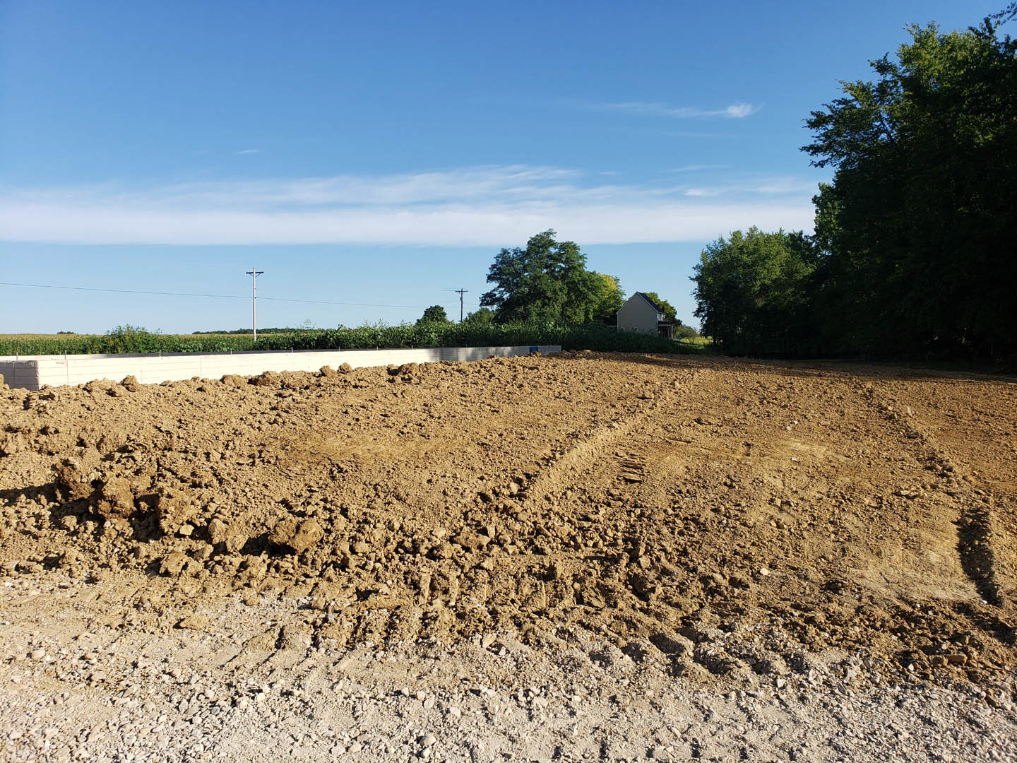 Dirt field with tire tracks bordered by leafy trees under a blue sky with scattered clouds, pile of soil near a fence in the foreground
