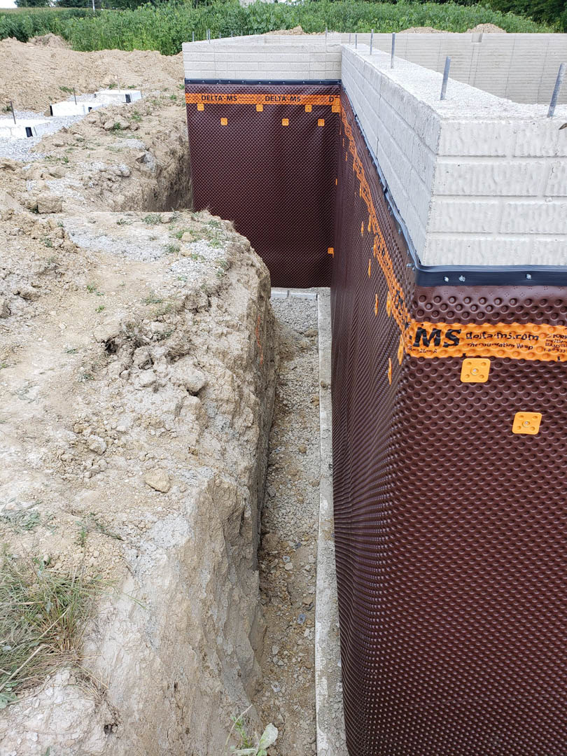 Concrete house foundation with orange foam insulation panels, surrounded by dirt and grass, waste container visible in background
