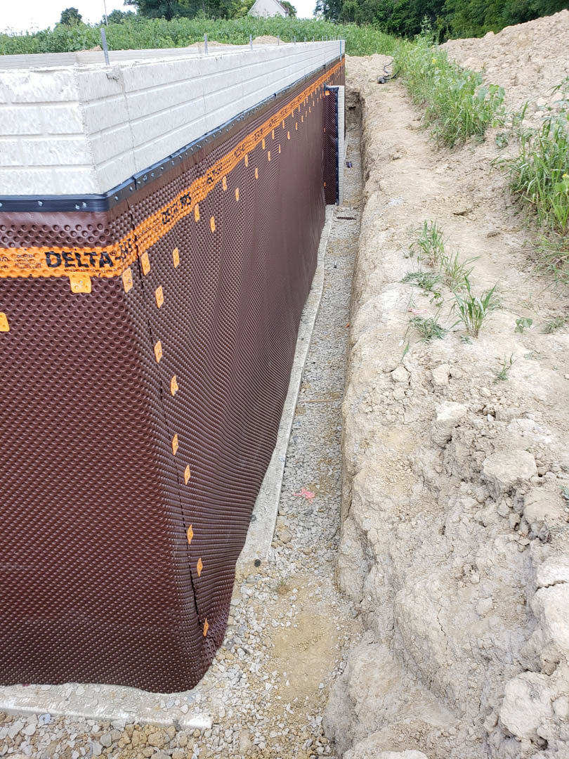 Brown wall with orange tape, black plastic sheeting partially covering the surface, dirt ground and sparse grass in foreground, fenced area with bushes and trees in background.