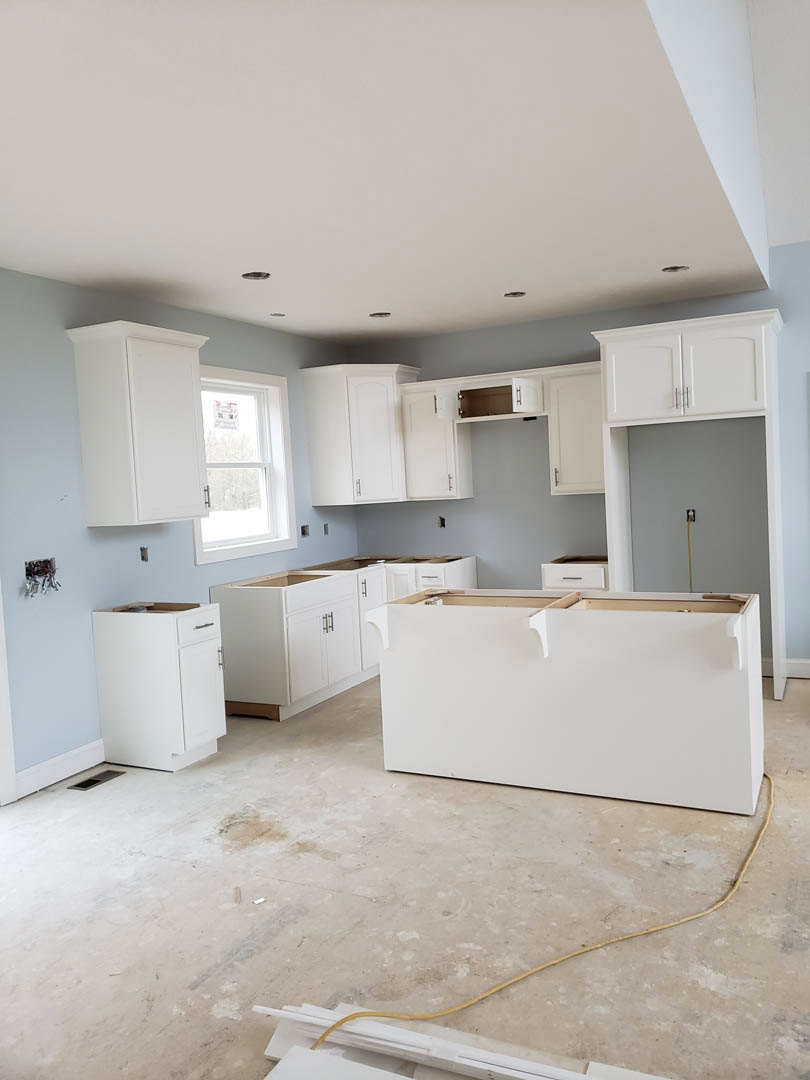 White kitchen cabinets with shaker doors, a window above the sink, concrete floor with yellow wire, unfinished walls and ceiling.