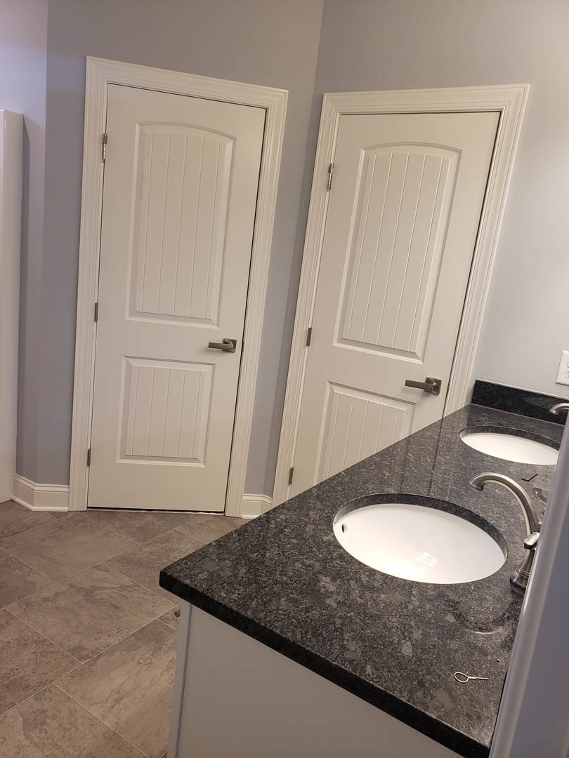 Bathroom with dual sinks, white cabinetry, two white doors with silver handles, large wall mirror, black circular ceiling light, and light-colored tile flooring