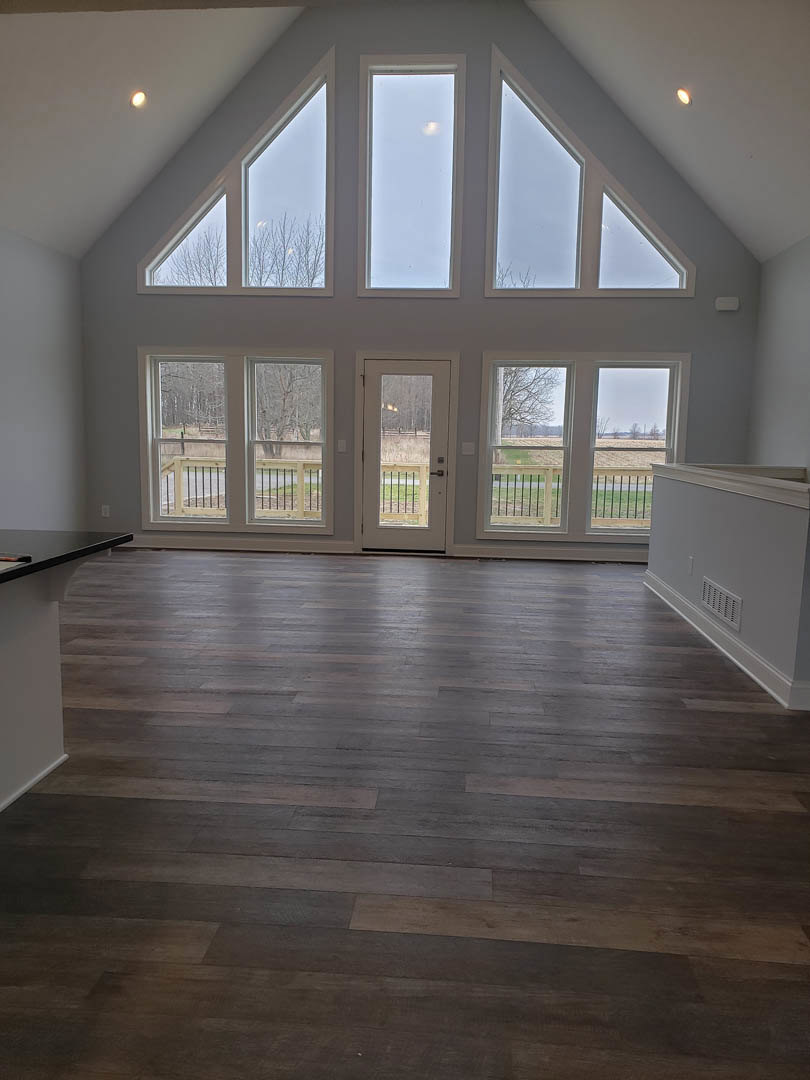 Hardwood floor room with large windows, white walls, and a door opening to a fenced grassy yard; sunlight reflecting on glass, wall vent visible